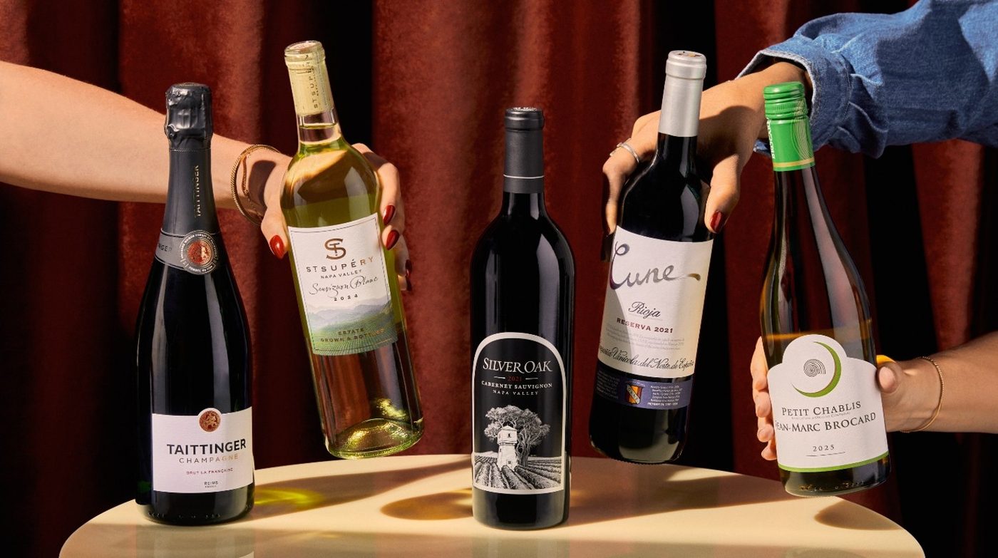 Hands holding bottles from Delta's new Delta One wine collection, including Champagne Taittinger, St. Supery Sauvignon Blanc, Silver Oak Cabernet Sauvignon, Cune Rioja, and Jean-Marc Brocard Petit Chablis